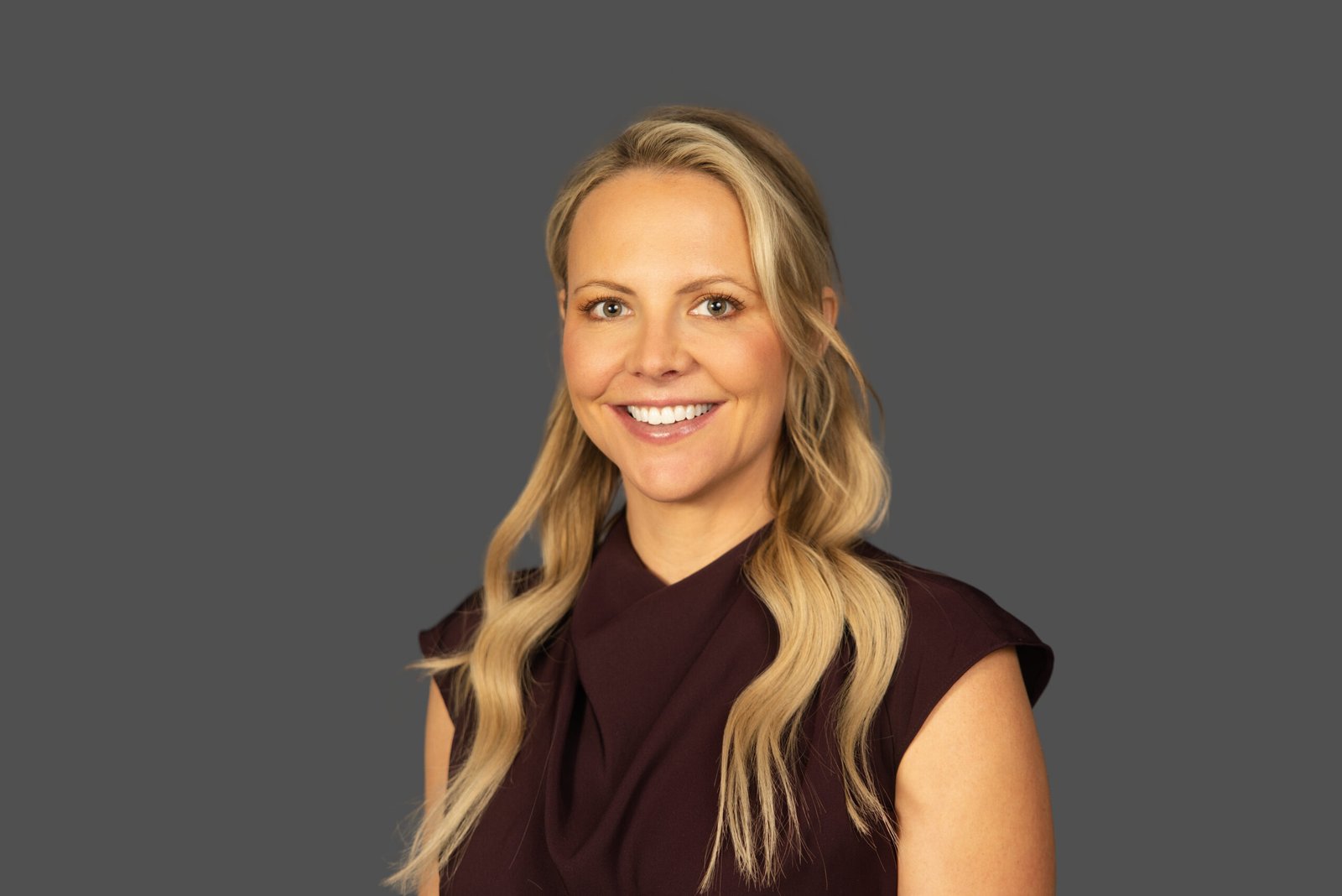 Smiling woman with long blonde hair in a maroon top against a plain gray background; conveys friendliness and professionalism.