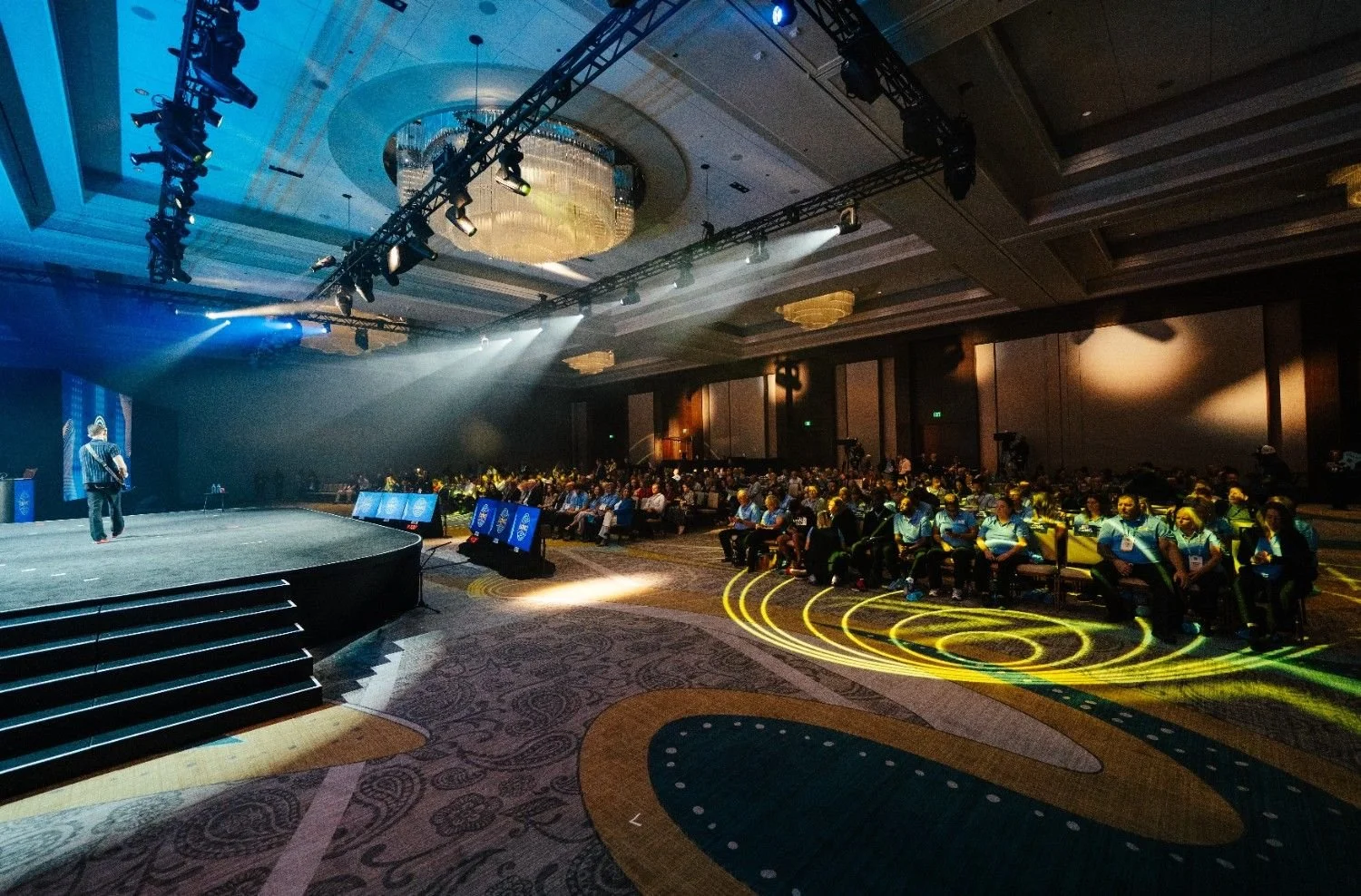 Full conference ballroom with a speaker on stage and camera equipment covering the audience