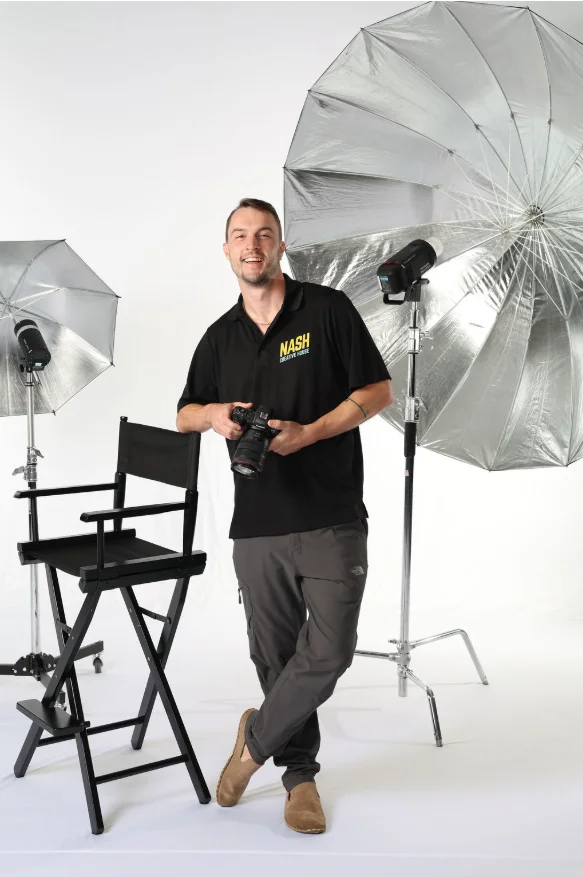 Nash Creative House photographer posing with a camera and studio lighting umbrellas in front of a director's chair